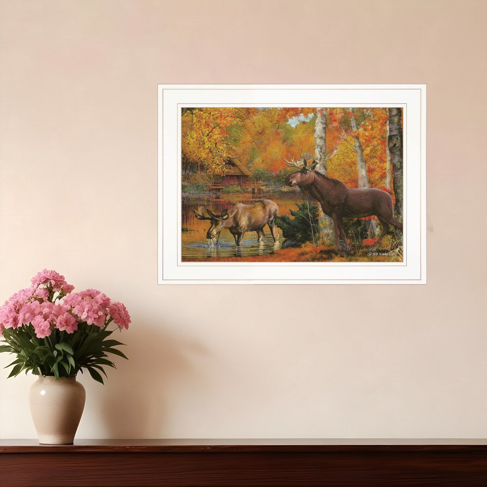 Framed painting of two moose in a forest scene on a wall above a vase with pink flowers.