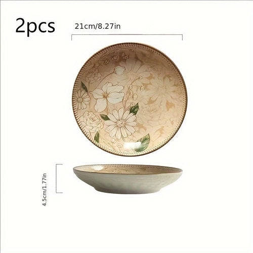 Two round ceramic plates with a floral design, displayed on a wooden table with a person's hands partially visible, holding one of the plates.