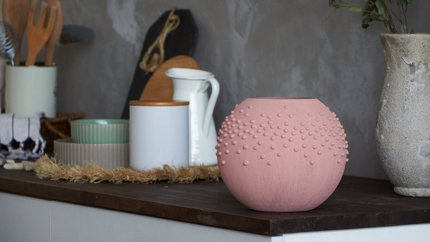 Pink textured vase on a shelf with kitchen utensils and decor items.