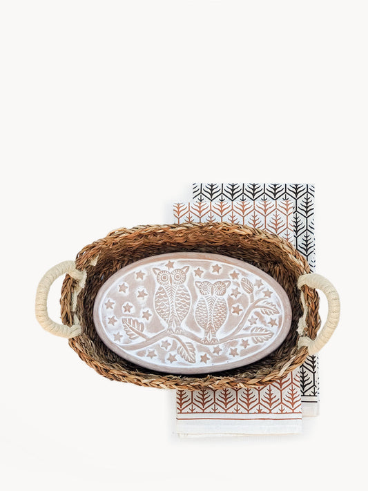 A terracotta bread warmer with an owl pattern, placed inside an oval seagrass basket, with a white and brown patterned tea towel.