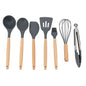 A set of seven kitchen tools with beech wood handles, including a whisk, basting brush, solid spoon, spatula, slotted turner, ladle, and tongs.