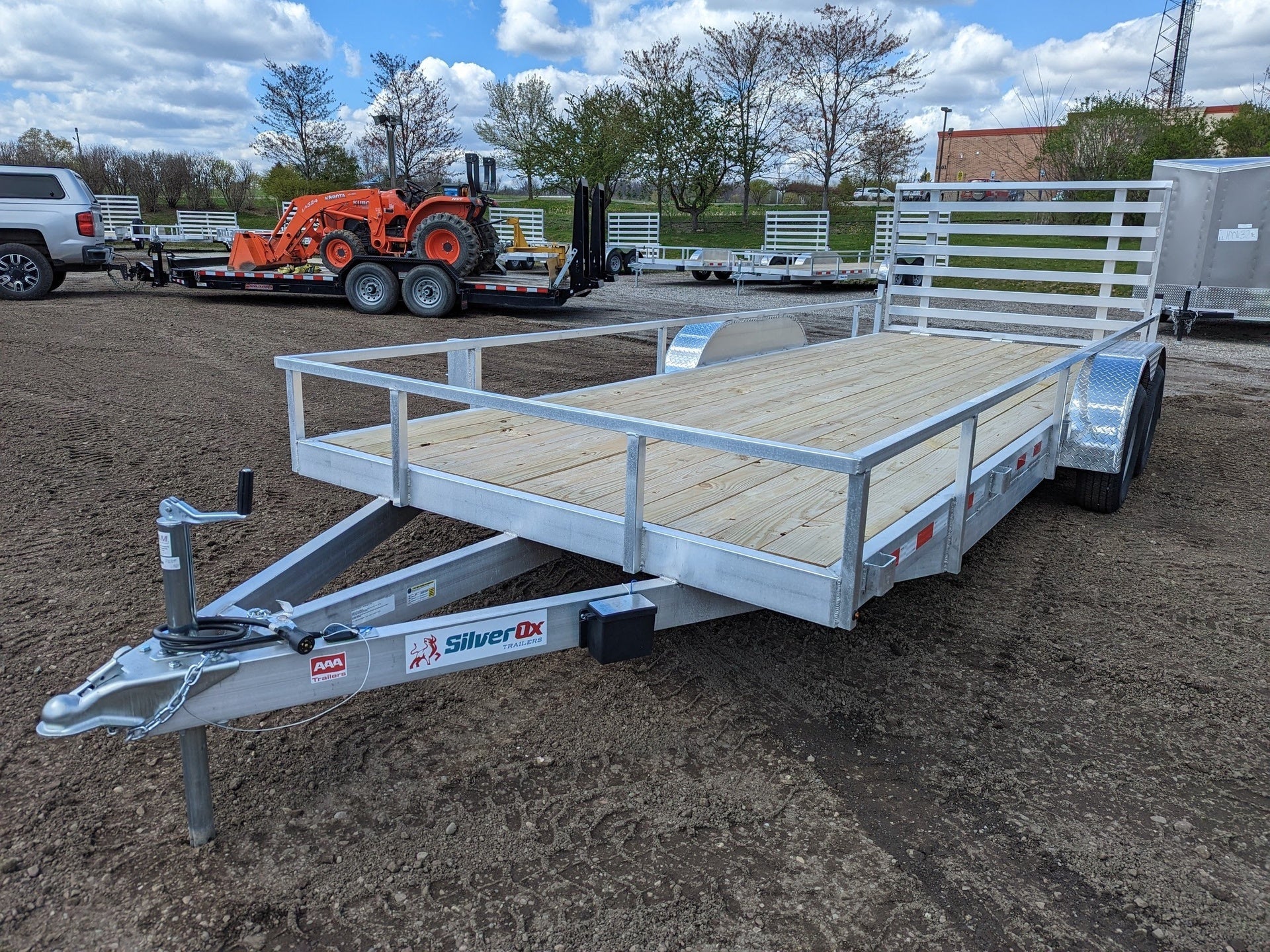 Silver utility trailer with wooden floor on a dirt lot with other trailers and equipment in the background.