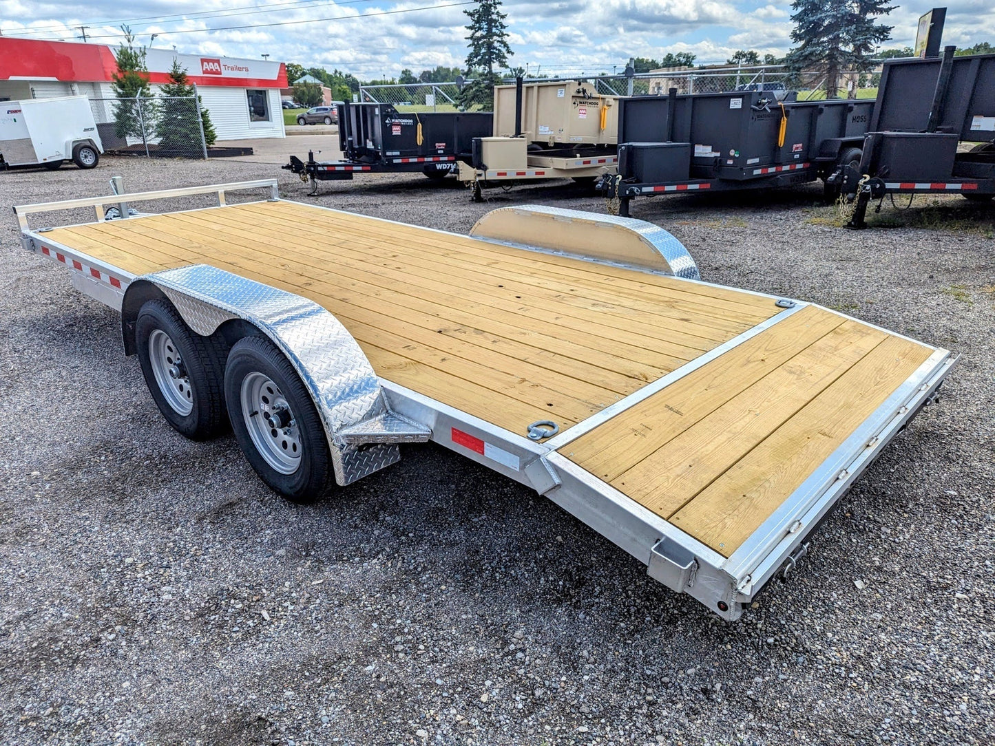 Wooden flatbed trailer on a gravel surface with a building and trees in the background.