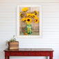 Framed artwork of sunflowers in a vase on a white wall above a red wooden table.