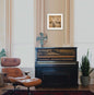 Piano in a room with a brown leather chair, fan, and framed picture on the wall.