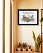 Framed artwork with chickens and text on a wall above a shelf with plants.