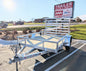 White utility trailer parked in a lot with a 'Trailer Sales & Parts' sign in the background.