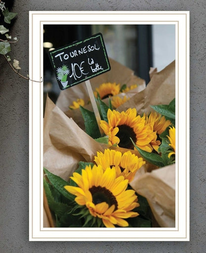 Framed picture of sunflowers with a small sign on a gray wall.
