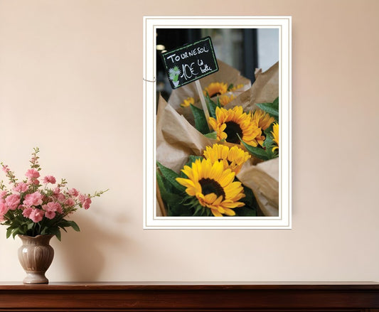 Framed picture of sunflowers on a wall with a vase of pink flowers below.