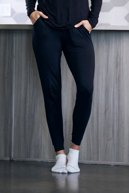 Person wearing black jogger pants and white socks standing against a gray wall.