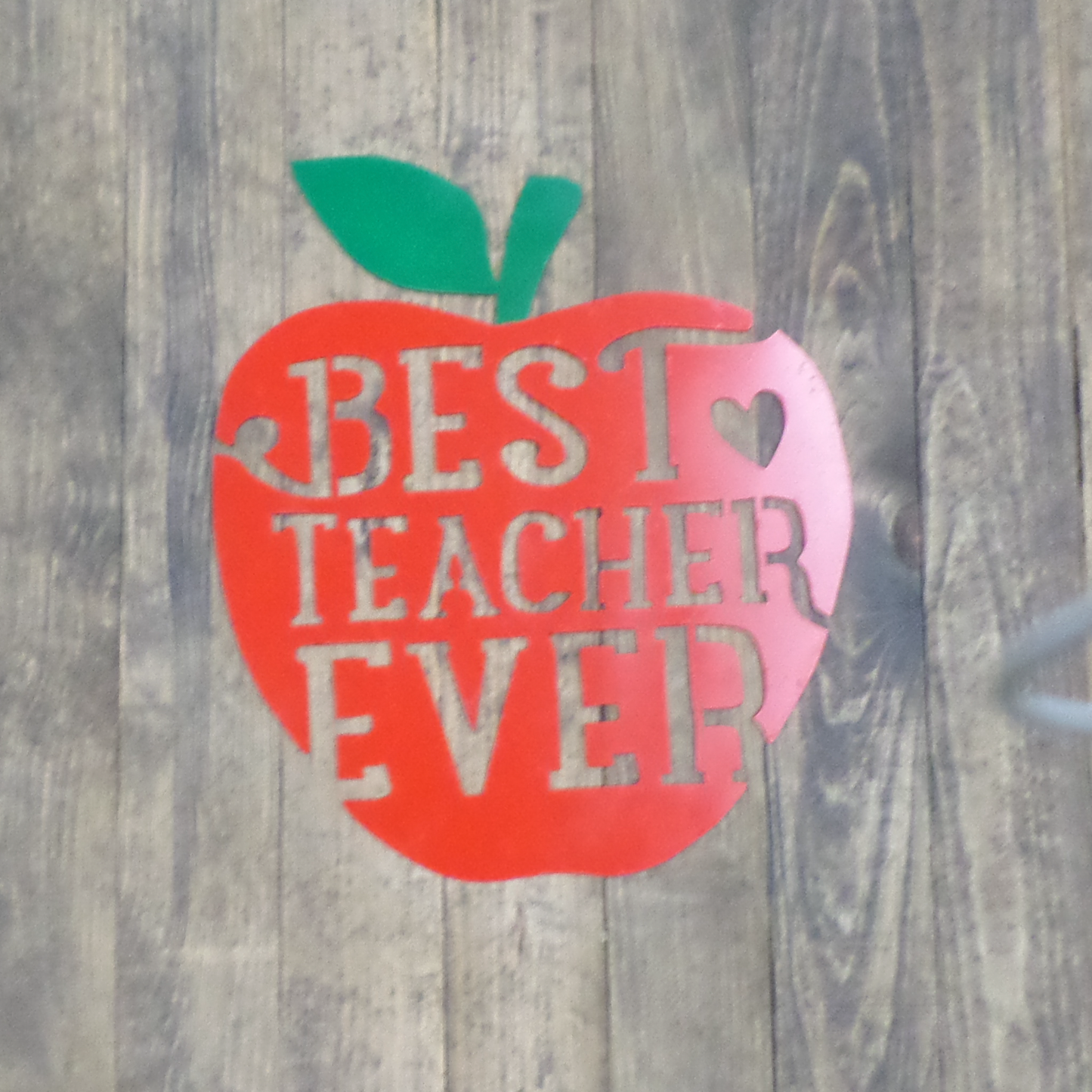 Decorative metal sign shaped like an apple with 'BEST TEACHER EVER' text on a wooden background