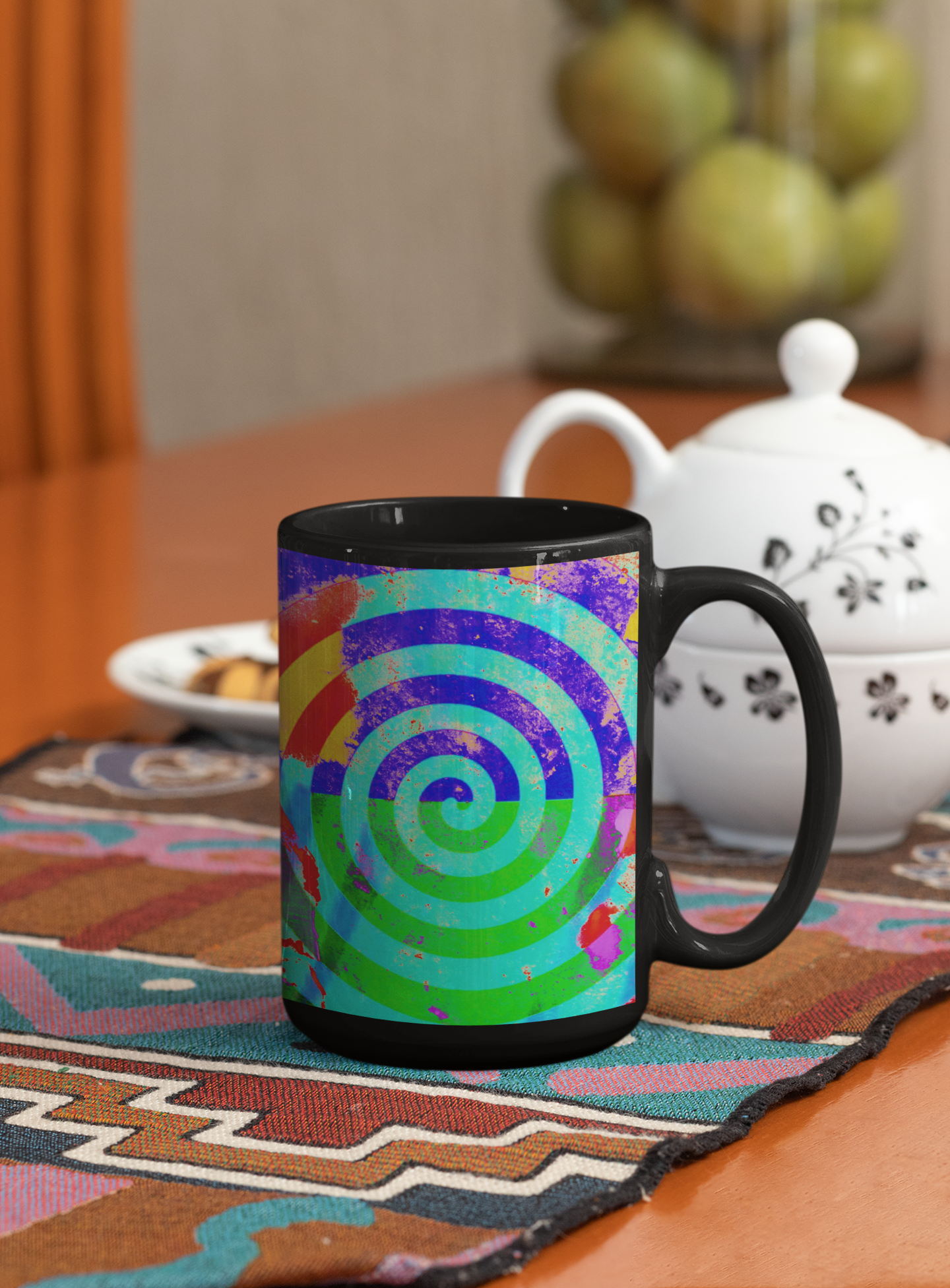 Colorful spiral-patterned mug with black handle on a table with a teapot and fruit bowl in the background.