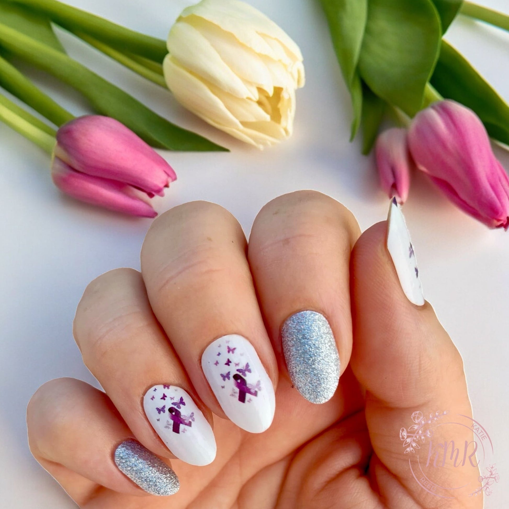 Nails with purple ribbon design and glittery accents in front of pink tulips