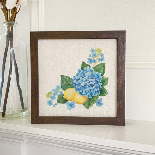 Framed artwork of blue flowers and lemons on a white surface with a vase in the background.