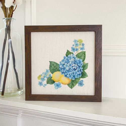 Framed artwork of blue flowers and lemons on a white surface with a vase in the background.