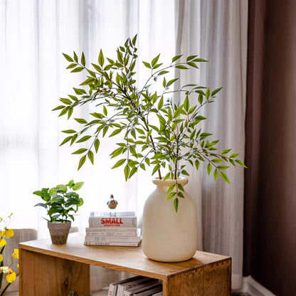 Vase with green leaves on a wooden table against a white curtain background