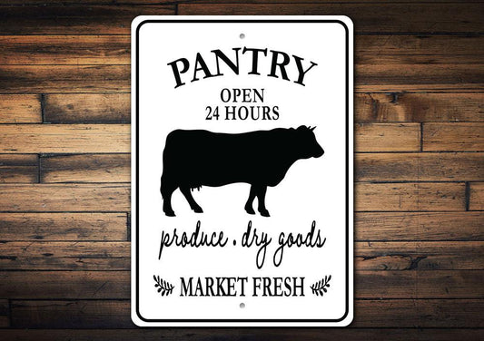 A white rectangular aluminum sign with black text and graphics, featuring the silhouette of a cow, and the words 'PANTRY OPEN 24 HOURS produce, dry goods MARKET FRESH', mounted on a wooden surface.