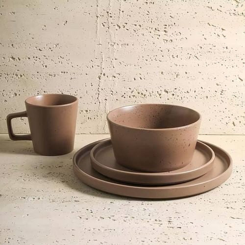 A set of matte brown stoneware dinnerware including a mug, a bowl, a plate, and a handled cup, displayed on a textured surface.