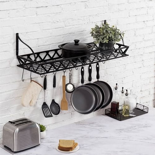 A black metal rectangular wall mounted kitchen pot rack with 12 hanging hooks, shown mounted on a white brick wall with various pots, pans, and kitchen utensils stored on and hanging from the rack.