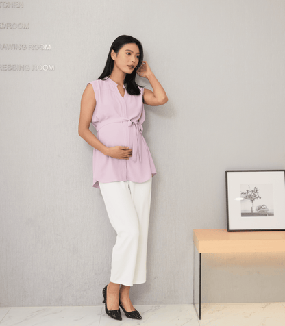 Woman wearing a pink sleeveless top and white pants standing against a gray wall.