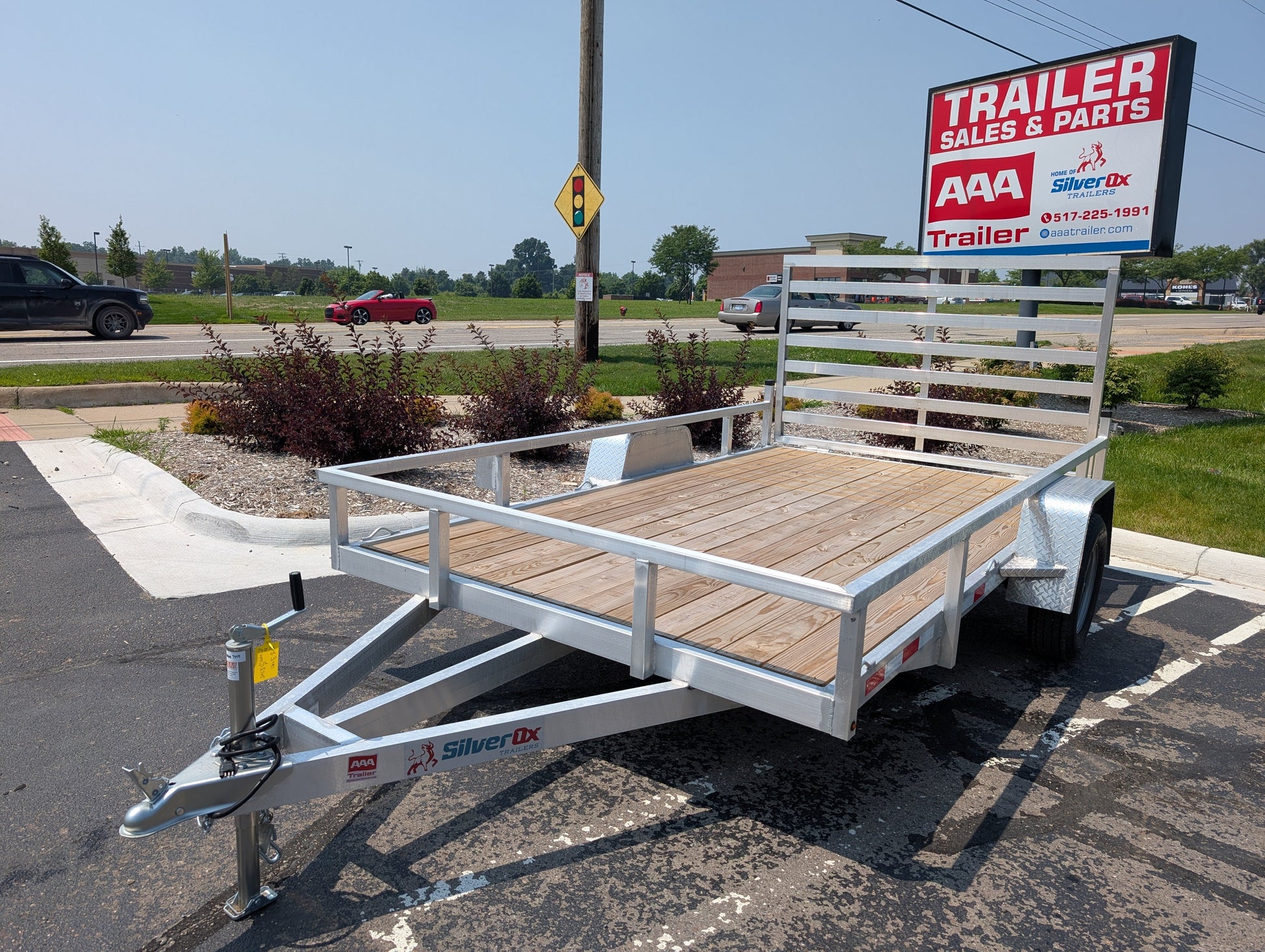 Silver trailer with wooden floor parked in a lot with a 'Trailer Sales & Parts' sign in the background.