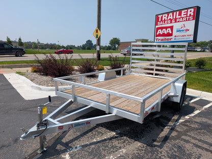 Silver trailer with wooden floor parked in a lot with a 'Trailer Sales & Parts' sign in the background.