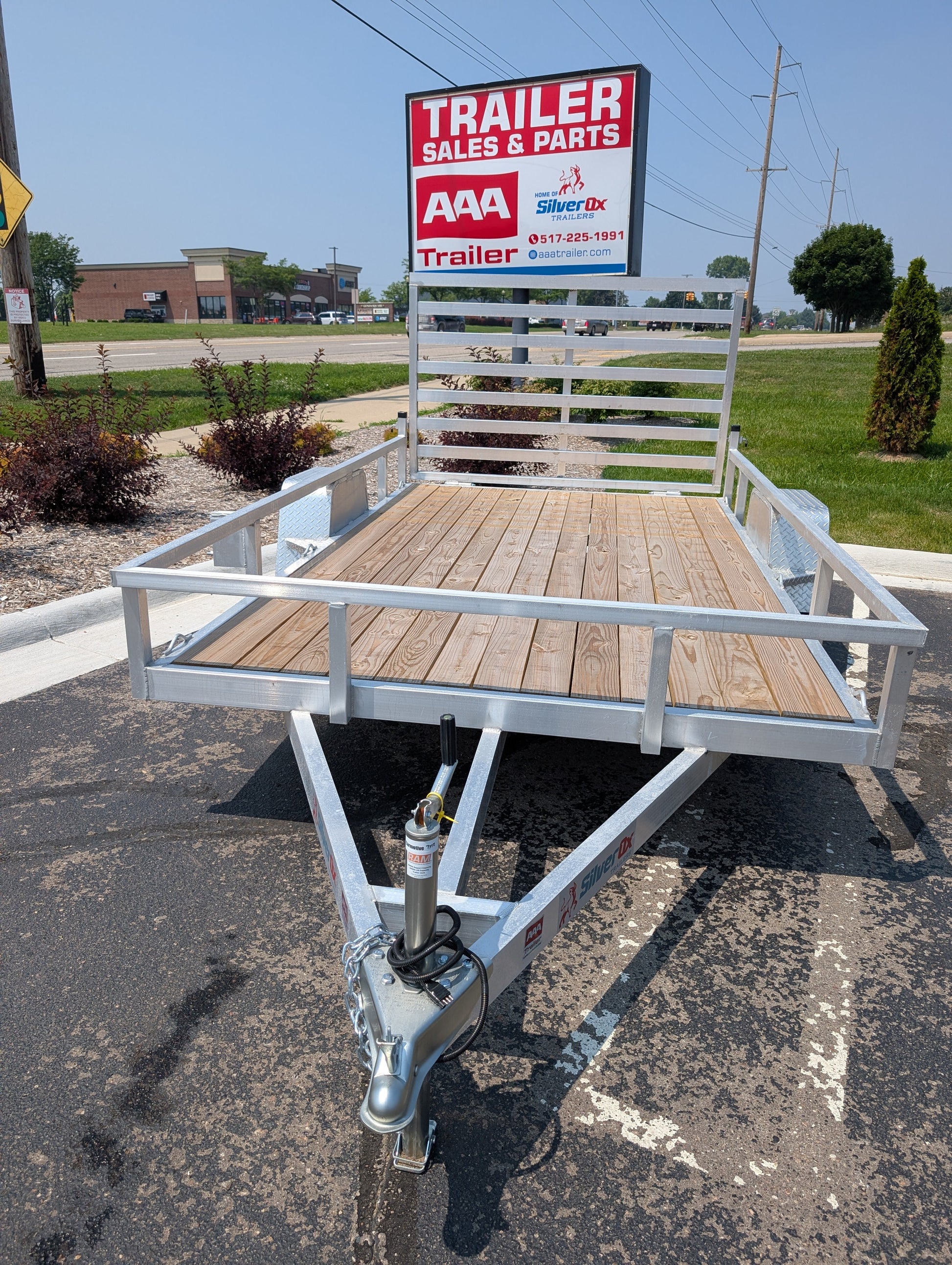 Small utility trailer with wooden floor on a paved surface, with a 'Trailer Sales & Parts' sign in the background.