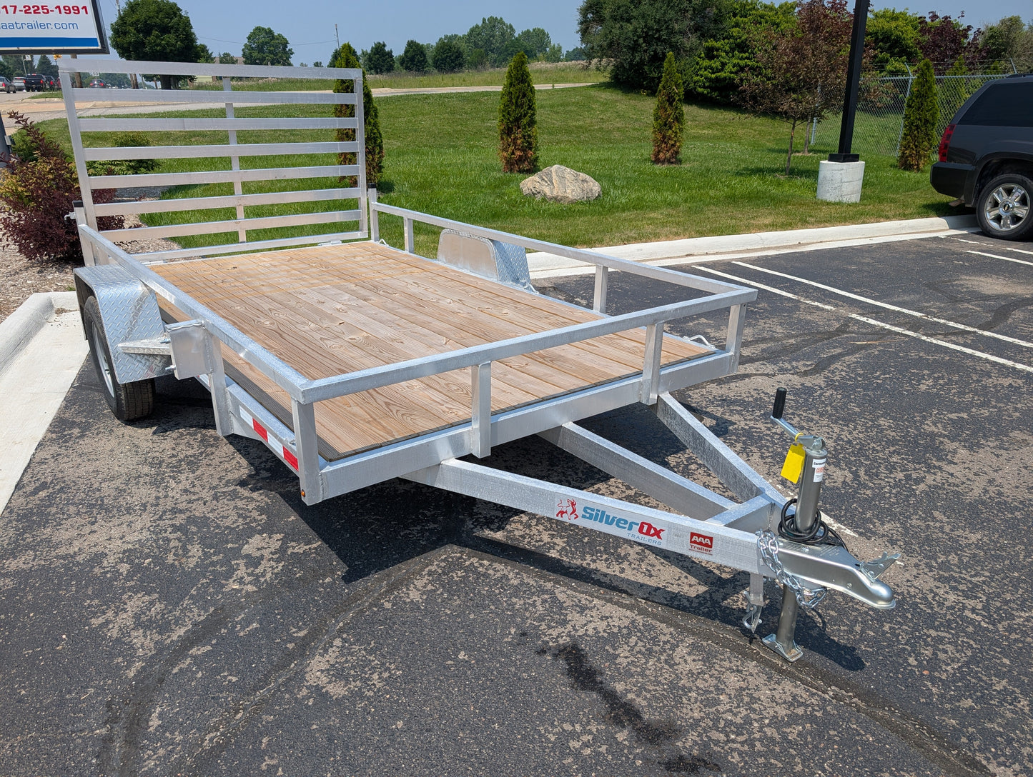 Silver Dx utility trailer parked on a paved surface with grass and trees in the background.