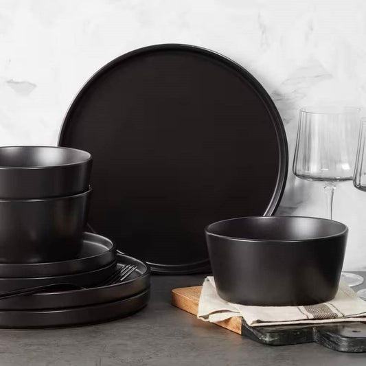 A 16-piece stoneware dinnerware set in matte black, including round dinner plates, salad plates, bowls, and mugs, displayed on a table with a neutral background.