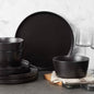 A 16-piece stoneware dinnerware set in matte black, including round dinner plates, salad plates, bowls, and mugs, displayed on a table with a neutral background.