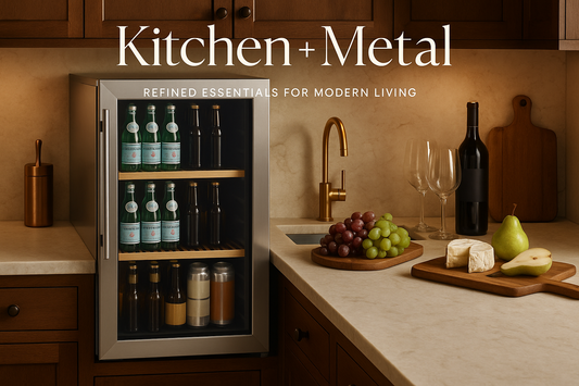A sophisticated marketing image showing a stainless steel beverage refrigerator in a warm, inviting kitchen setting. The refrigerator is styled with beverages and elegant food items on a marble countertop. The Kitchen + Metal logo appears in an elegant contemporary font with the tagline "Refined Essentials for Modern Living". The image beautifully balances cool metallics with warm wood tones and copper accents, creating a sophisticated yet welcoming atmosphere that embodies both elements of the brand name.