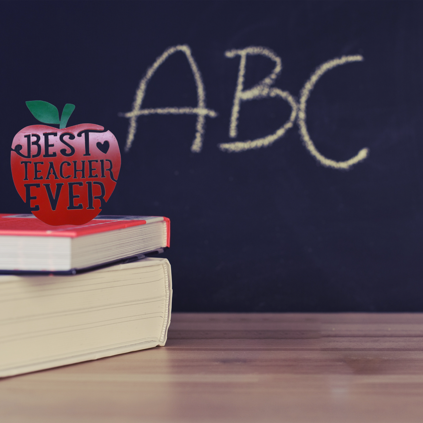 Stack of books with a decorative apple labeled 'Best Teacher Ever' in front of a blackboard with 'ABC' written on it.