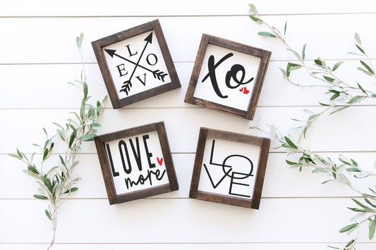 Four wooden frames with 'LOVE' and heart designs on a white wooden surface with greenery.