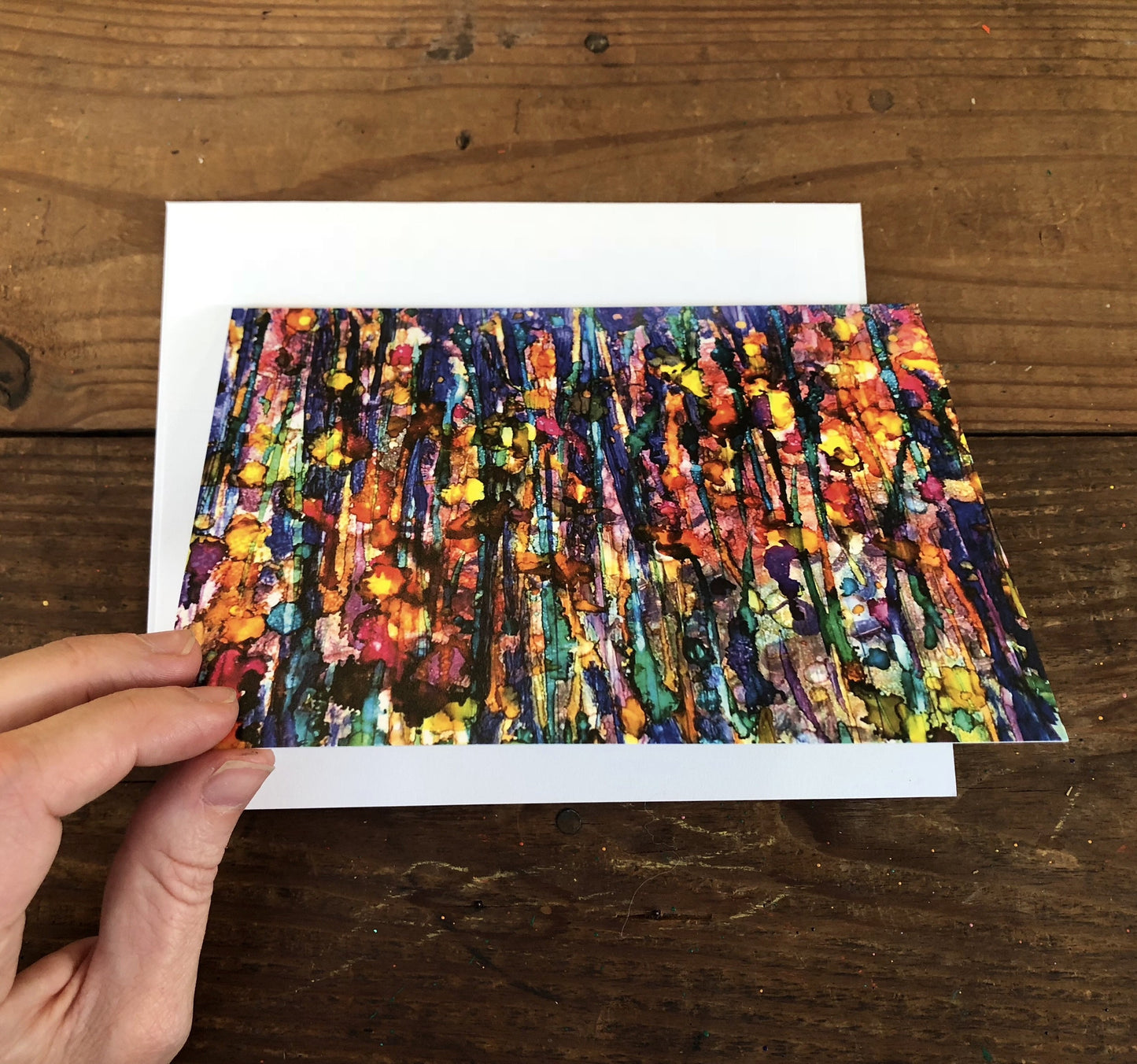 Colorful abstract painting on a white card held by a hand on a wooden surface