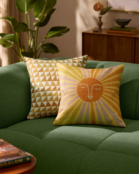 Two decorative pillows on a green couch with a plant and wooden cabinet in the background.