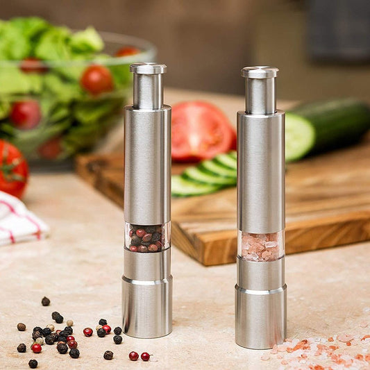 Two stainless steel salt or pepper grinders with visible spices inside, one filled with whole peppercorns. They are placed on a kitchen counter with vegetables in the background.