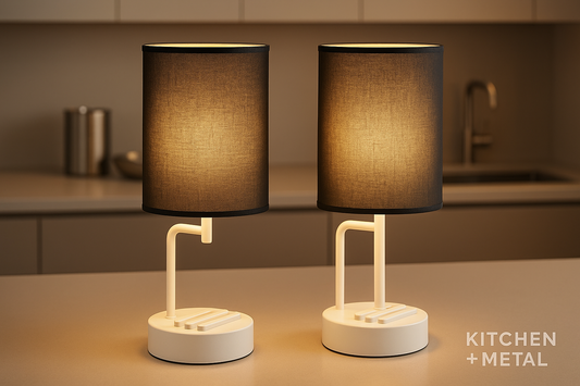 Two elegant black fabric shade cordless table lamps with white bases displayed in a modern kitchen setting. The lamps cast a warm, inviting glow against a stylish backdrop that features subtle metallic accents. The Kitchen + Metal branding appears discreetly in the corner, emphasizing the brand's sophisticated blend of functional lighting and premium kitchen aesthetics.